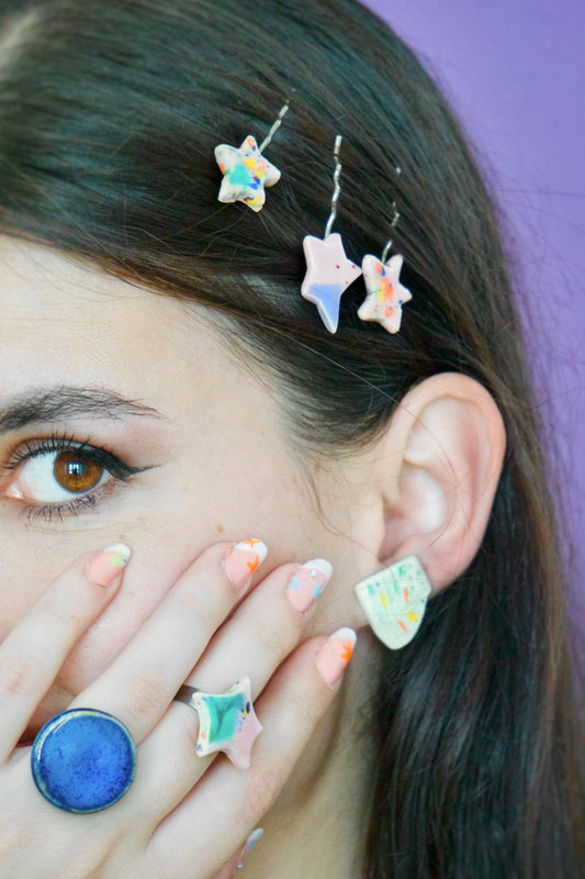 Cercel handmade din ceramică cu formă geometrică neregulată și glazură crem pal cu accente colorate, purtat la ureche, alături de inele ceramice colorate – bijuterii Ecluze Studio.