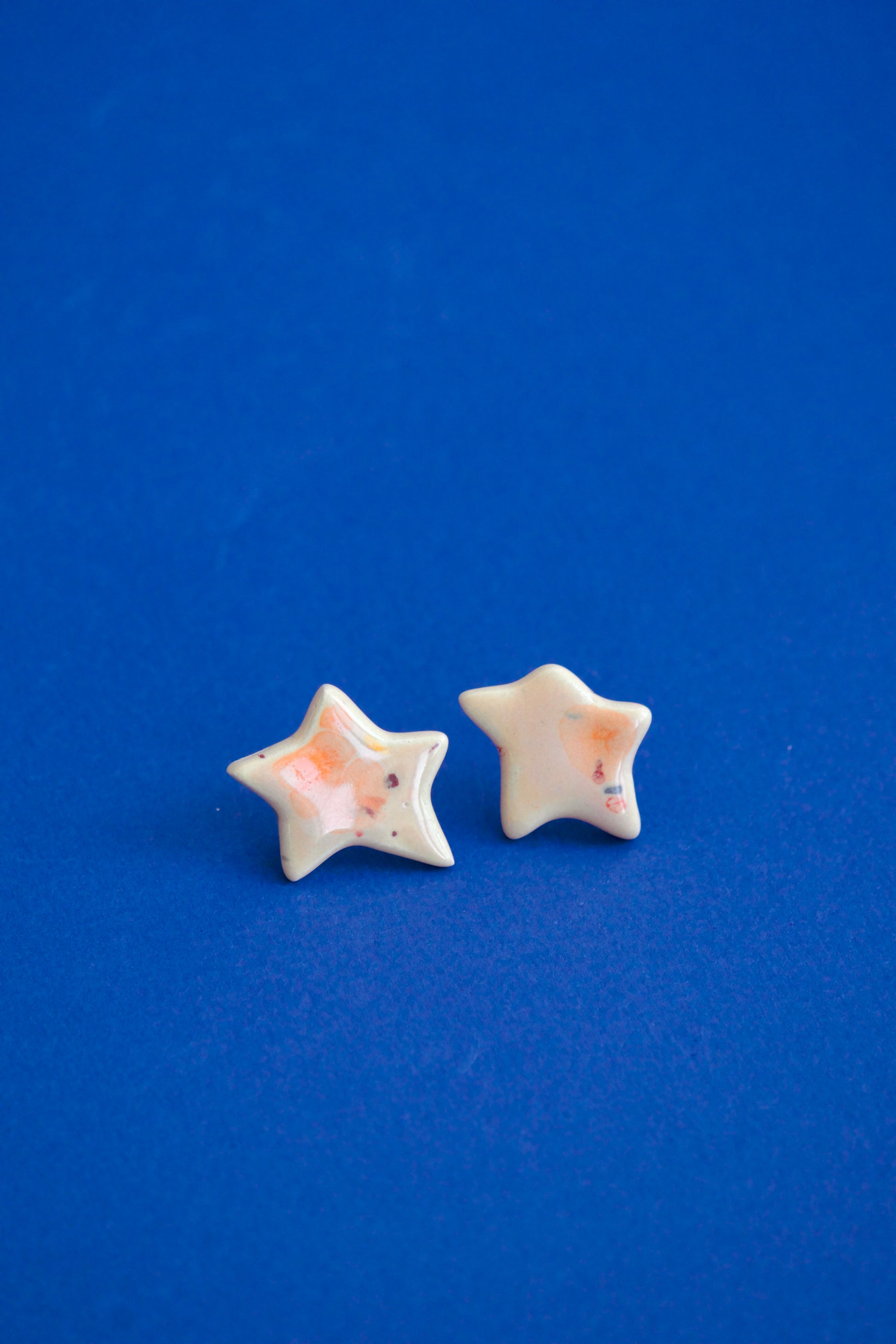 Cercei handmade din ceramică în formă de steluță Coral & Rosé, cu glazuri lucioase și tije hipoalergenice pe fundal albastru.