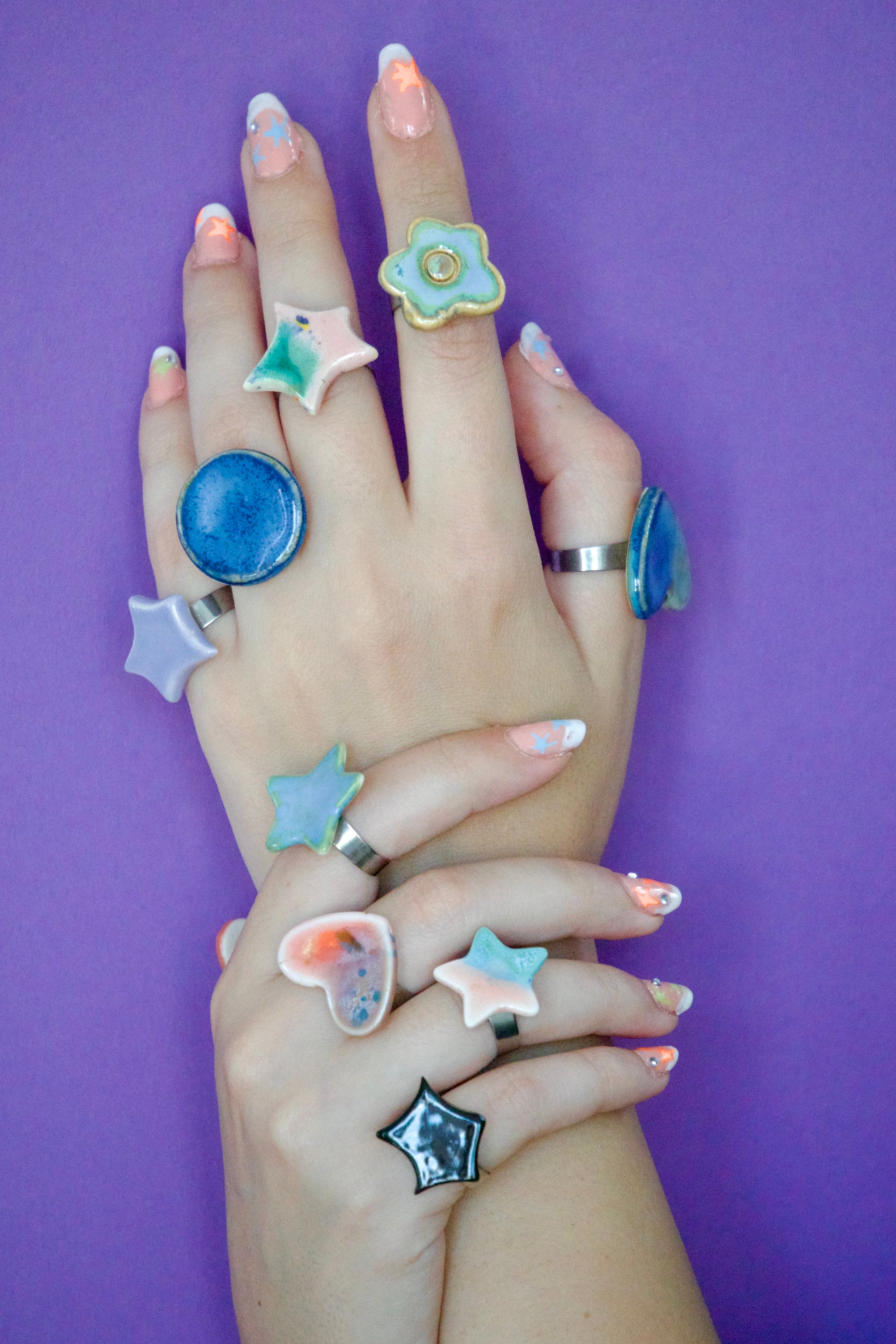 Mâini decorate cu multiple inele handmade din ceramică, în forme organice și geometrice (stea, inimă, floare, cerc), glazurate în nuanțe pastel și montate pe suport metalic reglabil – bijuterii Ecluze Studio.