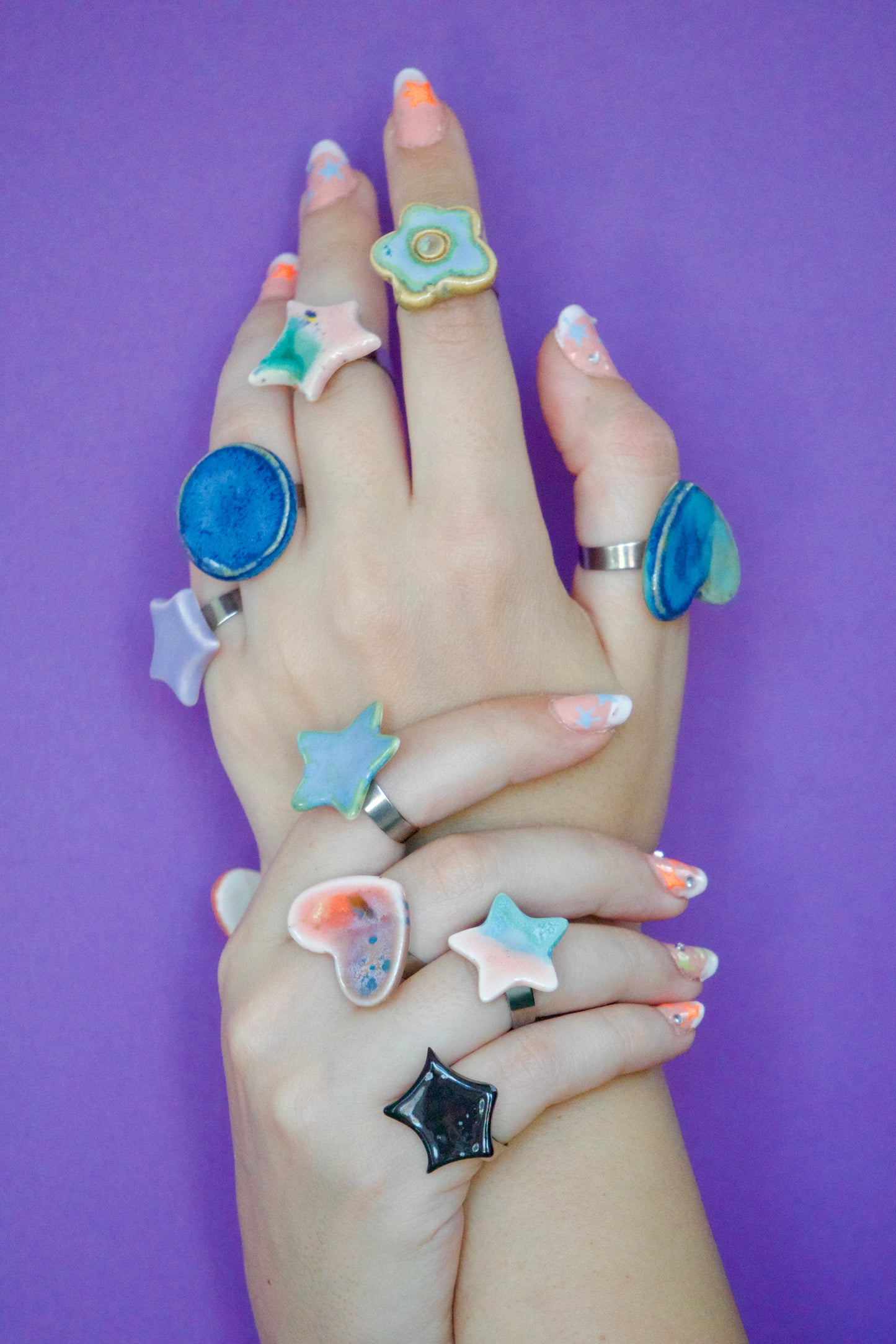Mâini decorate cu multiple inele handmade din ceramică, în forme organice (stea, inimă, floare, cerc), glazurate în nuanțe pastel și montate pe suport metalic reglabil pe fundal mov– bijuterii Ecluze Studio