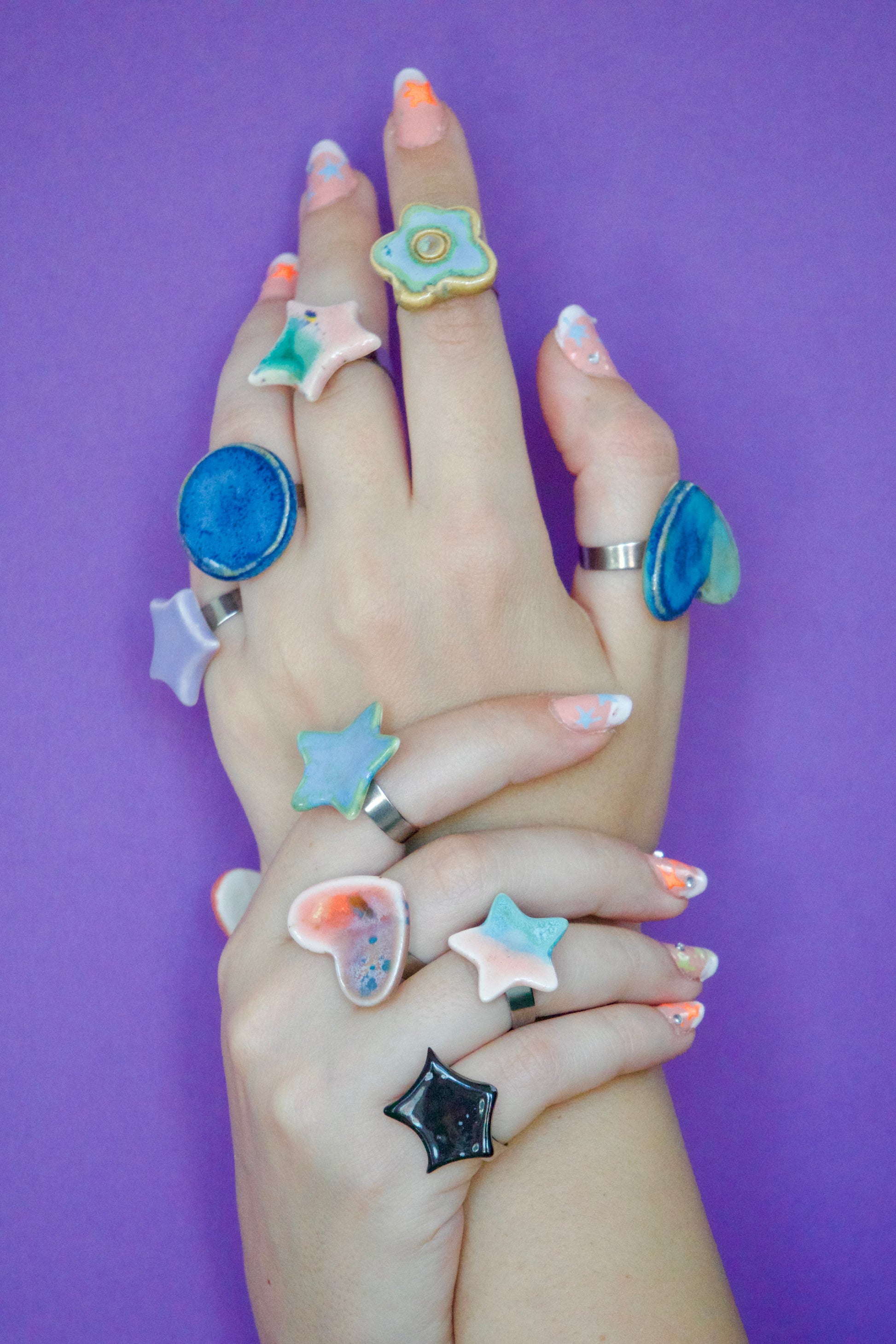 Mâini decorate cu multiple inele handmade din ceramică, în forme organice (stea, inimă, floare, cerc), glazurate în nuanțe pastel și montate pe suport metalic reglabil pe fundal mov– bijuterii Ecluze Studio