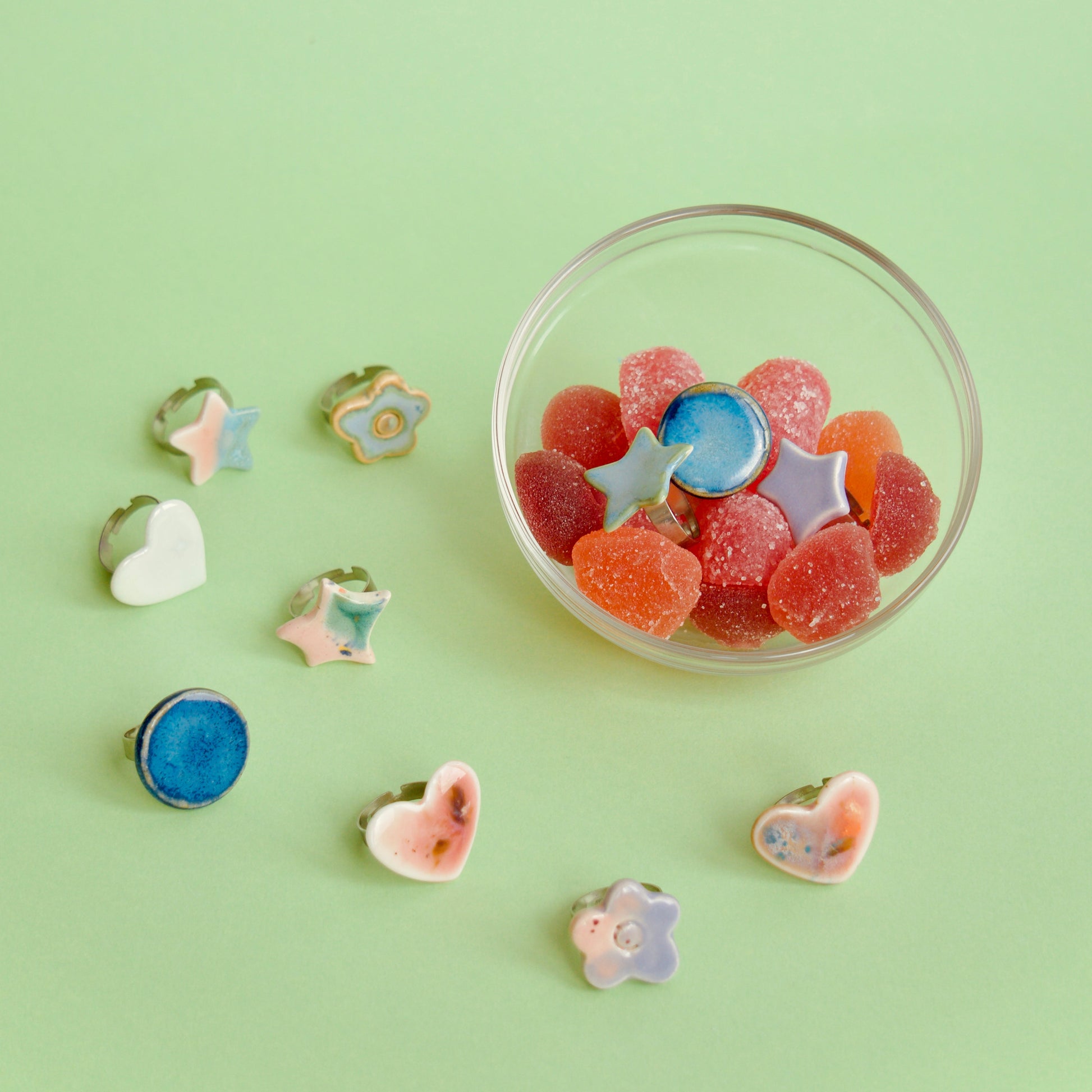 Colecție de inele handmade din ceramică în forme variate (stea, inimă, floare, cerc), glazurate în nuanțe pastel, alături de un bol cu jeleuri roșii și portocalii – bijuterii Ecluze Studio.