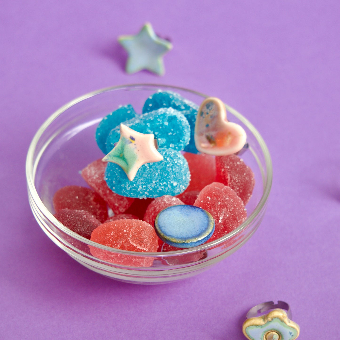 Bol din sticlă cu jeleuri roșii și albastre, decorat cu inele handmade din ceramică în forme organice și culori pastel, pe fundal mov – bijuterii Ecluze Studio.