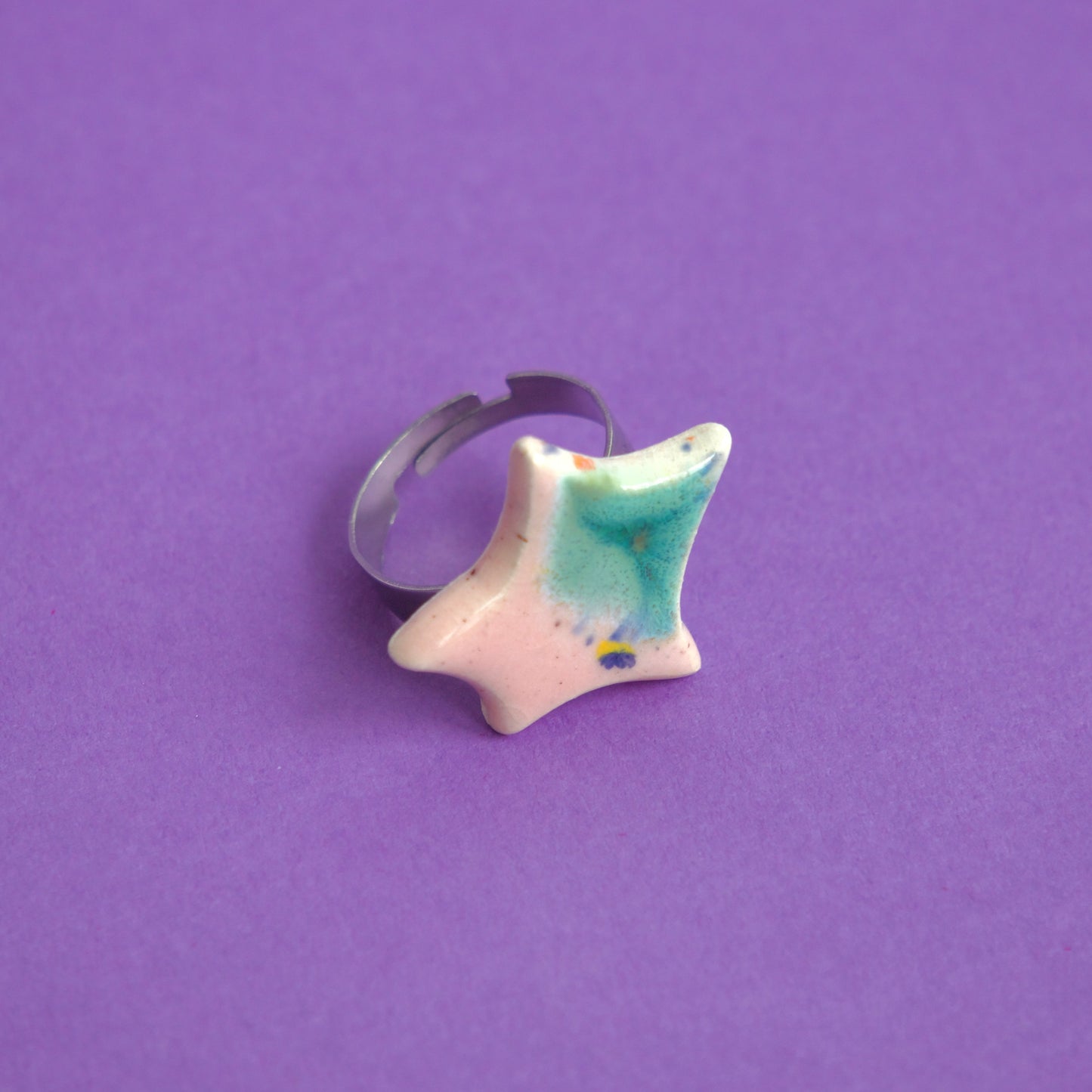 Inel handmade din ceramică cu formă organică, glazură roz și verde, montat pe suport reglabil din metal, pe fundal mov – bijuterie Ecluze Studio.
