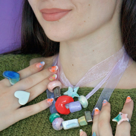 Colier handmade din ceramică cu mărgele colorate și panglică transparentă, purtat de o femeie, accesorizat cu inele ceramice în nuanțe pastel – bijuterii Ecluze Studio.