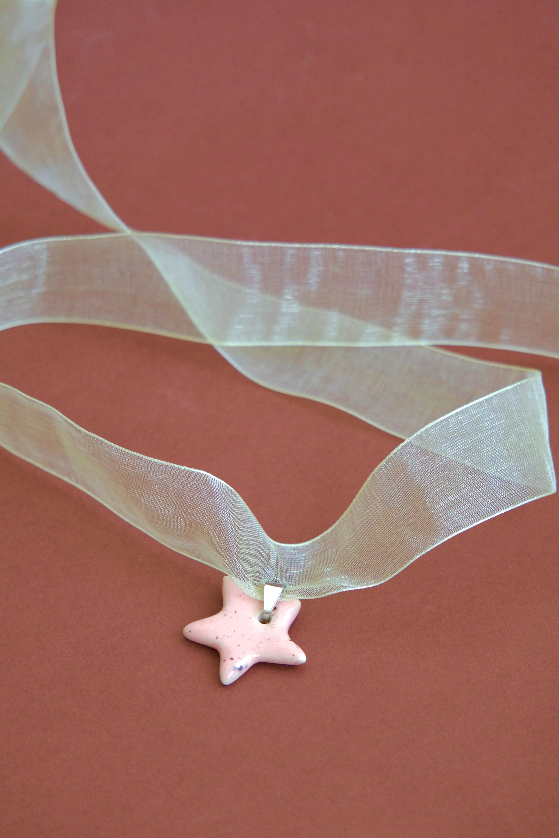 Colier unicat cu pandantiv stea roz-pastel din ceramica pe snur din organza galben deschis pe fundal maro.