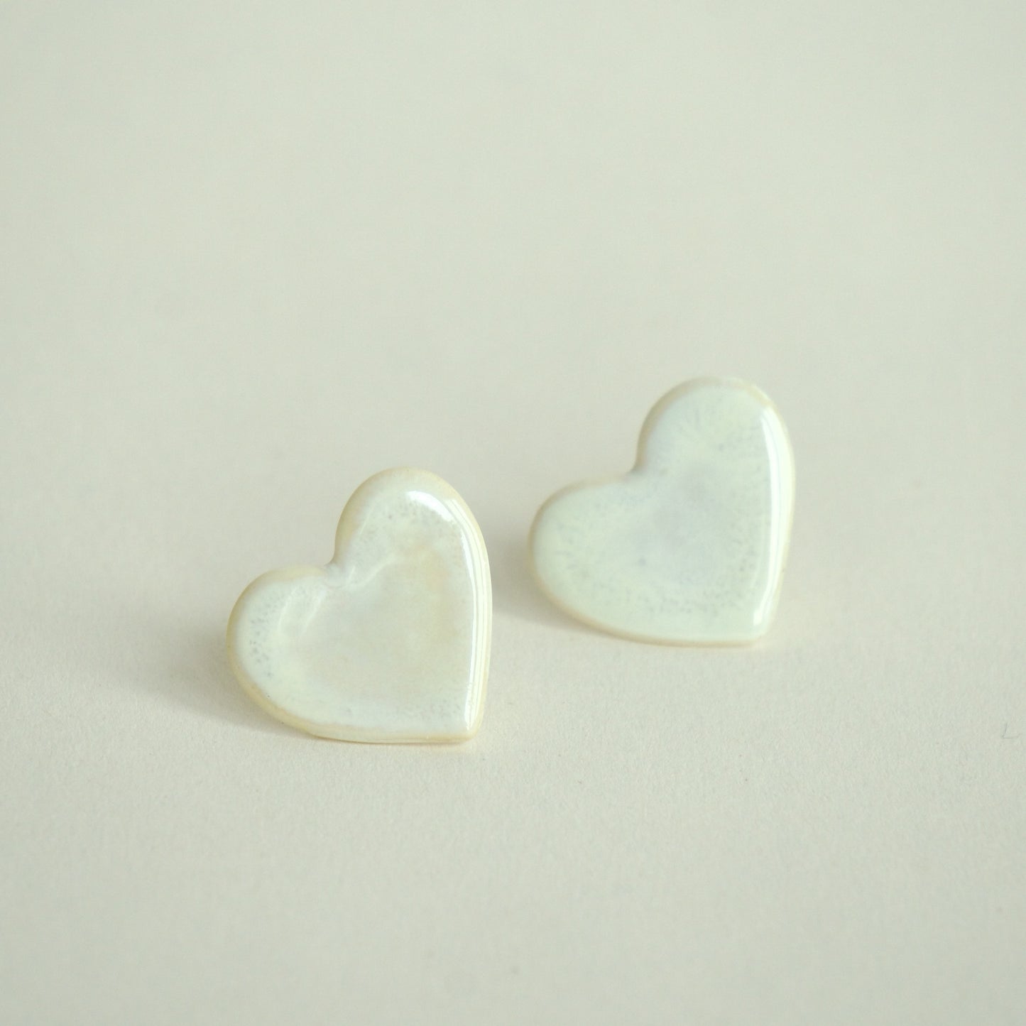 Cercei handmade din porțelan în formă de inimă, alb perlat,finisaj lucios pe fundal crem.