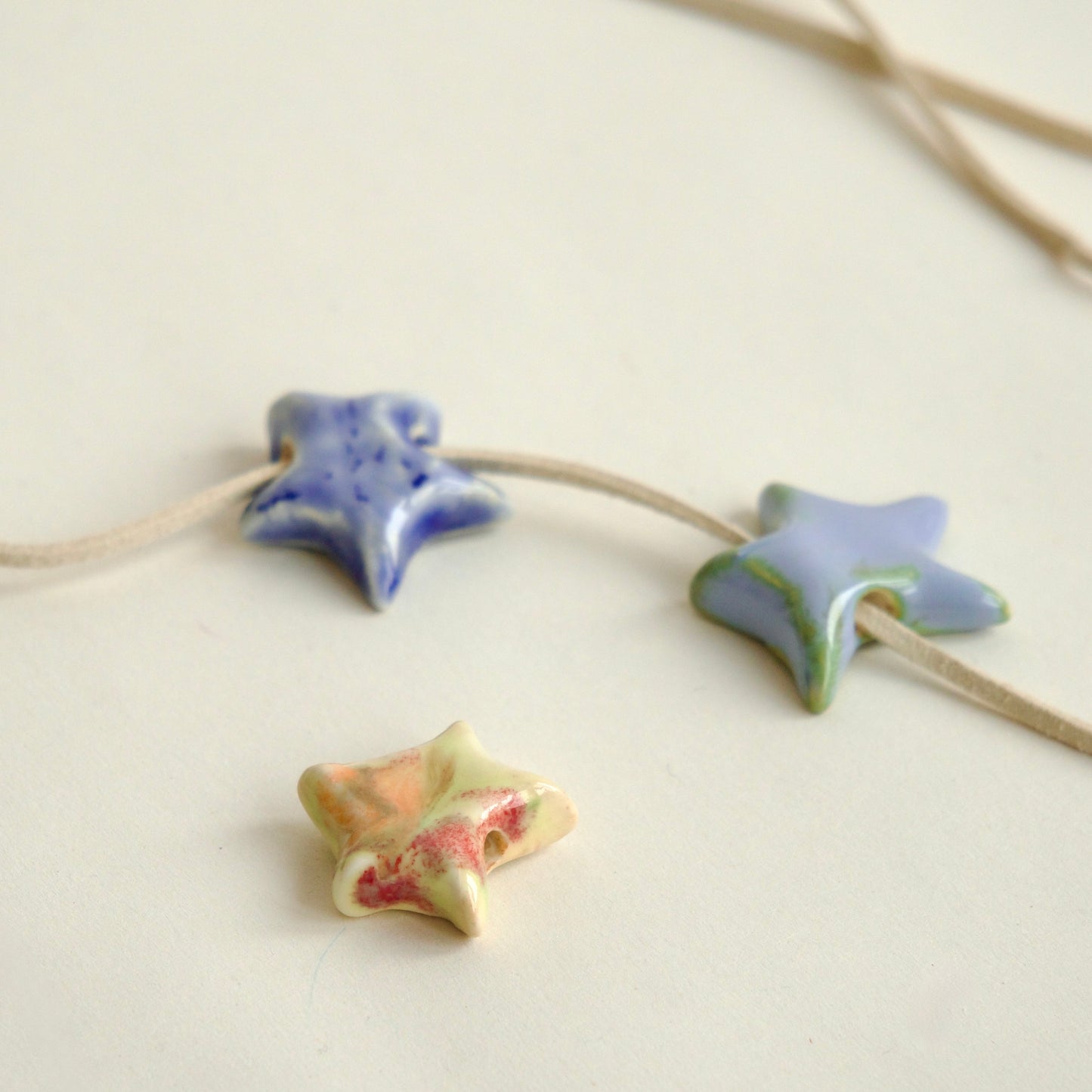 Charmuri handmade din ceramică în formă de steluță, culori variate – albastru, verde pastel, roz coral – pe șnur din suede bej.